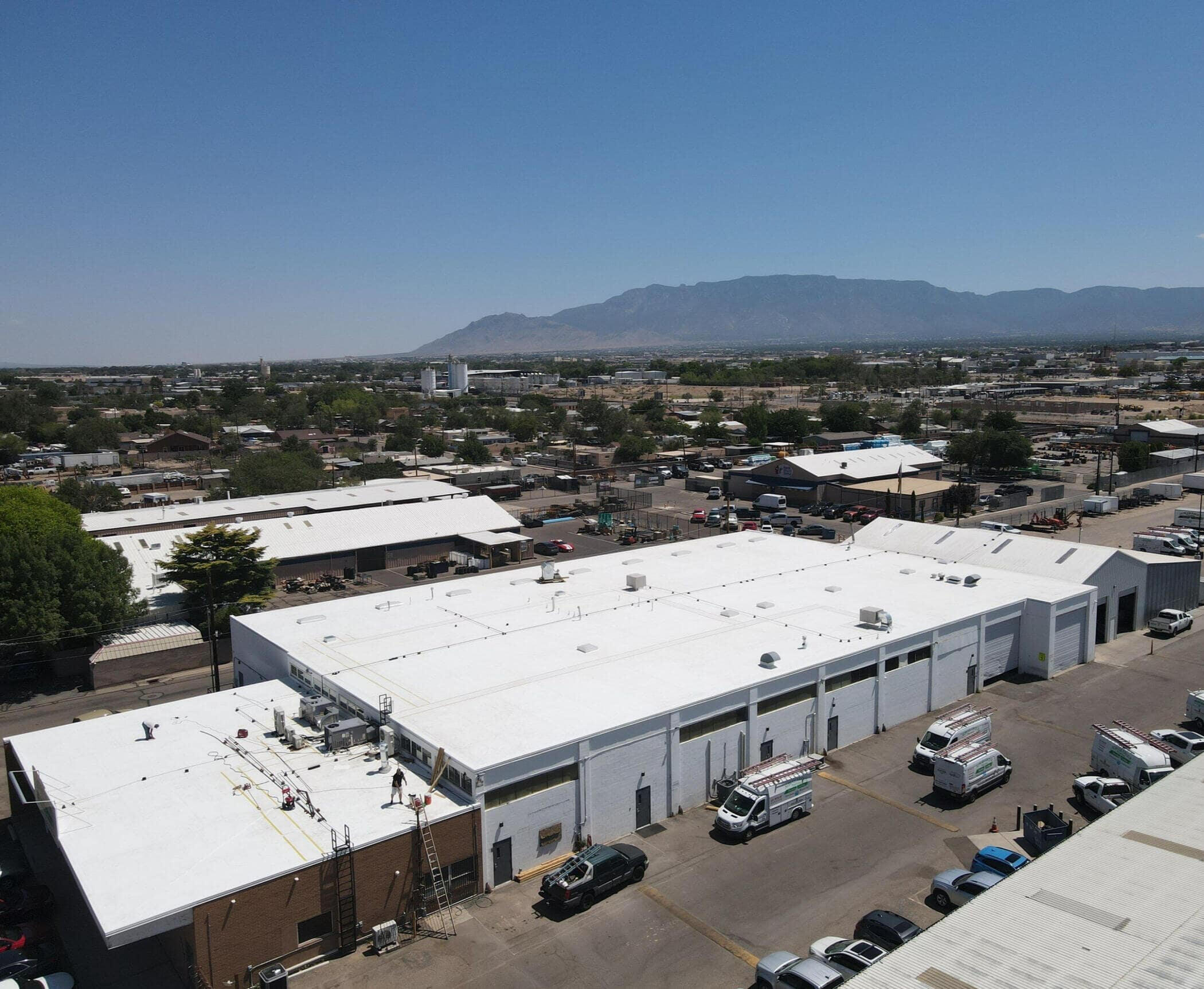 Aerial view captured by a drone of a large flat roof commercial building in Albuquerque, New Mexico. A ladder leans against the side of the building, and a roofer is seen working on the new white TPO roof.