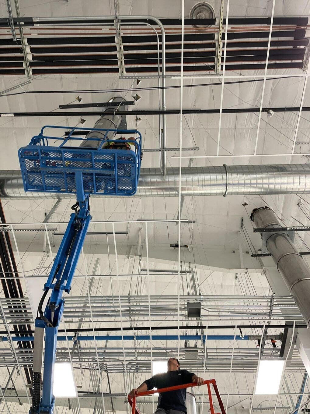 Roofers utilizing an aerial lift inside a commercial building in Albuquerque, New Mexico, to cut penetrations in the ceiling for vents that will connect to the roof.