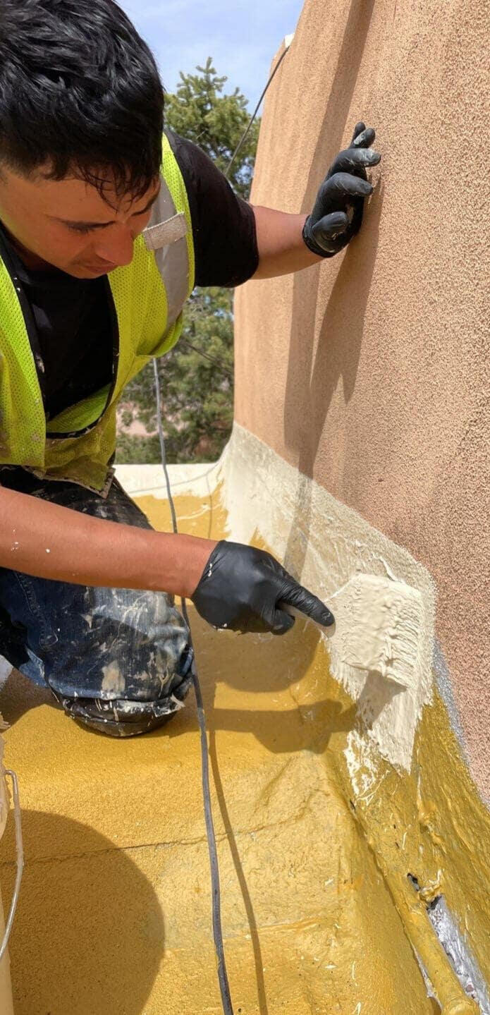 Roofer applying silicone mastic to the wall flashing of a modified bitumen flat roof system in Albuquerque, New Mexico, to repair the wall flashing before applying a silicone coating to fix a leak in the area.