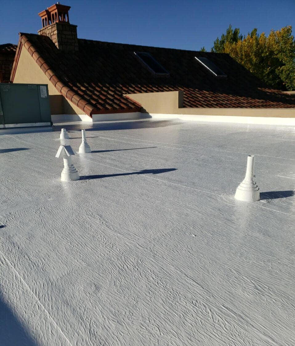 View on a well-maintained flat roof system in Albuquerque, New Mexico, featuring a white silicone coating with multiple pipe penetrations properly sealed. In the background, another section of well-maintained tiled roof is visible.