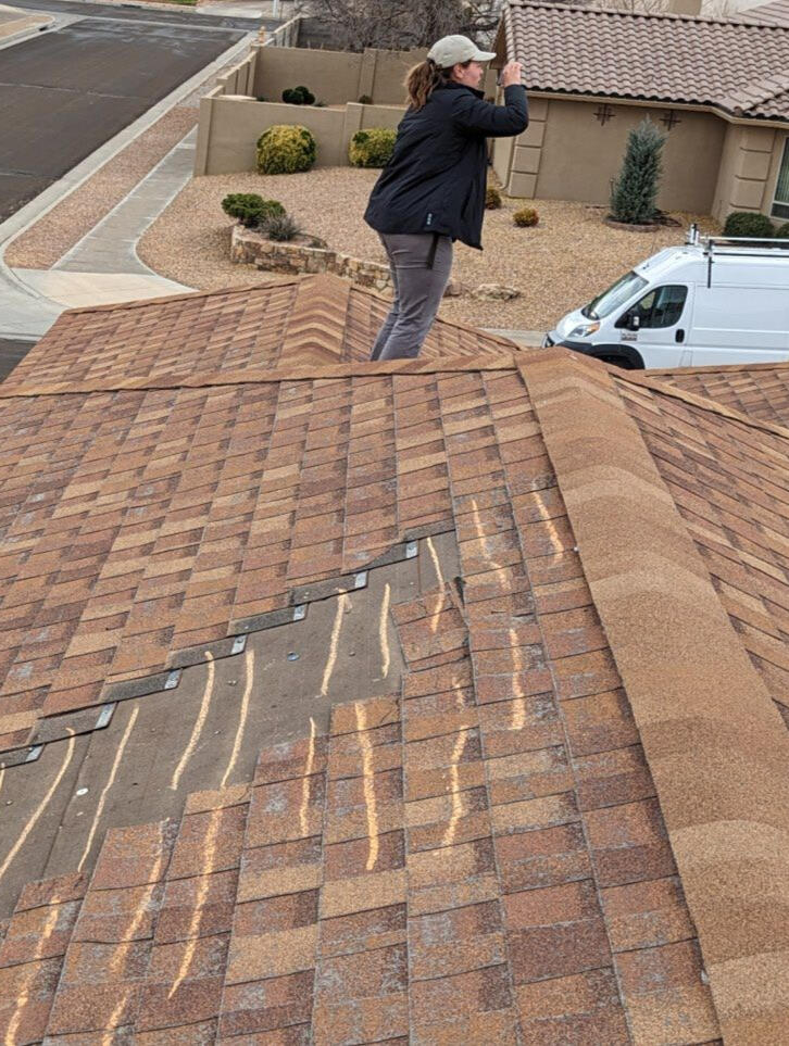 Female insurance adjuster inspecting wind damage on a pitched shingle roof for an insurance claim in Albuquerque, New Mexico. Missing shingles are marked with chalk to highlight the areas of concern.