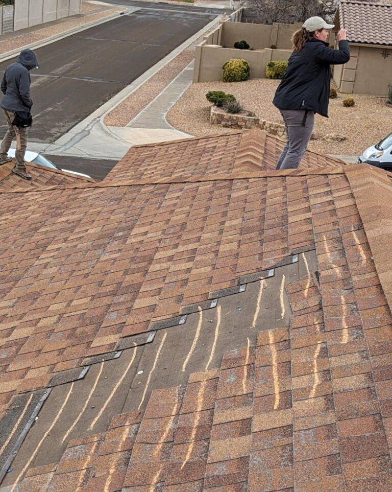 Two insurance adjusters on a shingle roof in Albuquerque, New Mexico, inspecting the roof for wind damage. Damaged shingles are visible, indicating areas of concern.